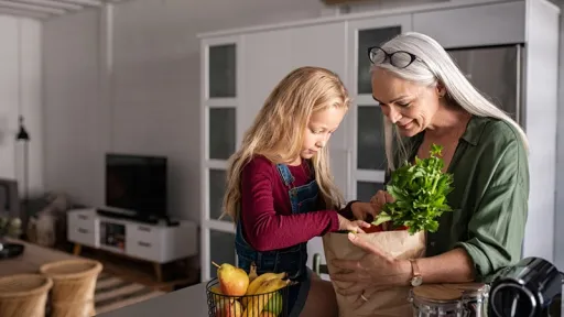 Grandmother and her granddaughter holding grocery shopping bag with vegetables at home. Happy old grandma holding paper bag with vegetables while grandchild removing food from bag. Senior lovely woman and little girl sitting on kitchen counter with groceries.
