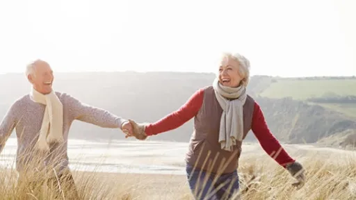 Senior Couple Walking Through Sand Dunes On Winter Beach Having Fun