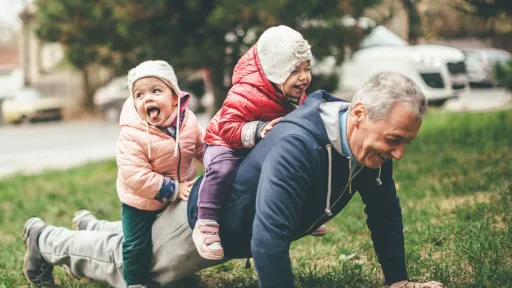 A photo of a playful grandfather and granddaughter. They are casually dressed and playing in the park. They exercise together. A grandfather is exercising while granddaughters are sitting on his back.