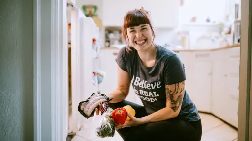 A smiling woman takes some vegetables and meat from the fridge for preparing a meal.
