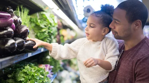 A pre-school age girl helps her dad pick out veggies in the produce section at the grocery store. He is holding her next to the produce and she is picking out eggplant.