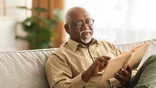 Senior African American Man Reading Book Sitting On Couch At Home, Wearing Eyeglasses. Retired Male Enjoying Reading New Novel Or Business Literature On Weekend. 